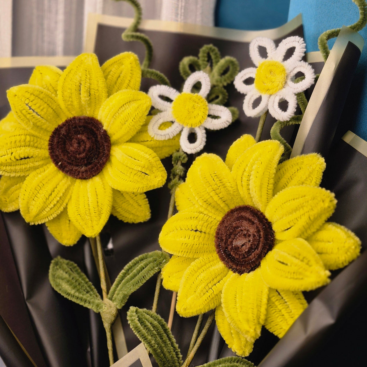 Three handmade crocheted flower bouquets made from pipe cleaners, featuring two large yellow sunflowers with dark brown centers and two smaller white daisies with yellow centers, wrapped in black paper.