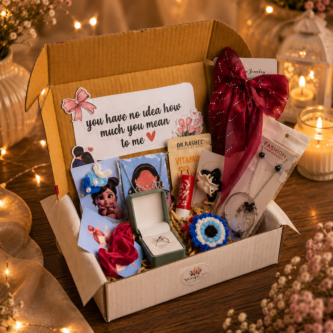 A curated gift box filled with assorted treats and surprises, photographed on a wooden table with warm string lights and candles in the background. The box contains a handwritten note, a red ribbon, a small ring box, a tube of lip balm, a packaged accessory, and other gift items.