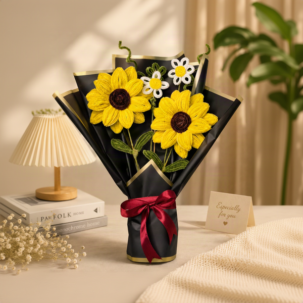 A handmade crochet sunflower bouquet wrapped in black paper with gold trim and a red ribbon, displayed on a tabletop with a small lamp, books, and a card in the background.