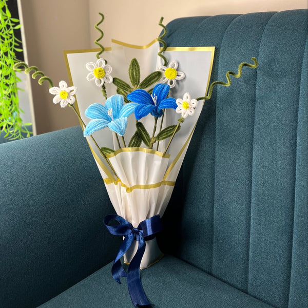 A small bouquet of artificial flowers wrapped in white paper with gold trim and tied with a blue ribbon, placed on a teal upholstered chair in an indoor setting.